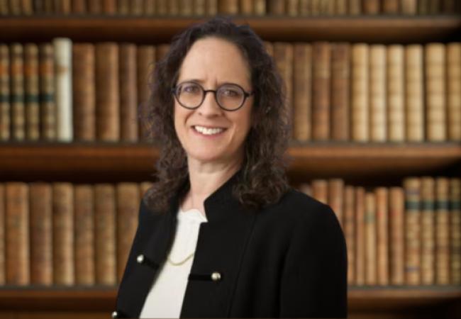 Alyza Lewin, glasses, black jacket, in front of bookshelf bg
