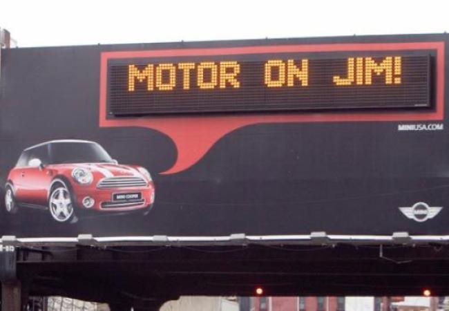 Mini Cooper billboard displaying a personalized message that says, "Motor on Jim!"