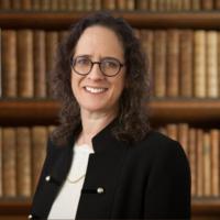 Alyza Lewin, glasses, black jacket, in front of bookshelf bg