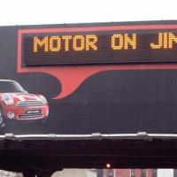 Mini Cooper billboard displaying a personalized message that says, "Motor on Jim!"